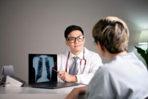 Kind and polite young Asian medical doctor orthopedic surgeon wearing stethoscope showing x-ray image from laptop while explaining and soothing patient about diagnosis and treatment plan in examination room in hospital. Asian qualified physician using laptop to discuss with patient on symptoms and health care and cure in medical clinic.