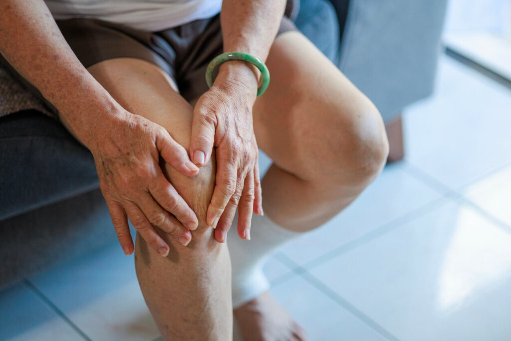 A senior Asian woman with osteoarthritis is massaging her knee while sitting on a sofa in the living room at hom